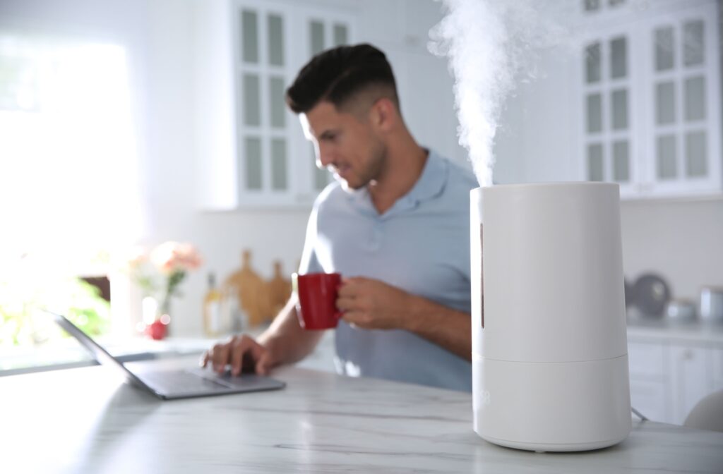 A humidifier is working in the foreground while an out-of-focus person in the background is working on a laptop.