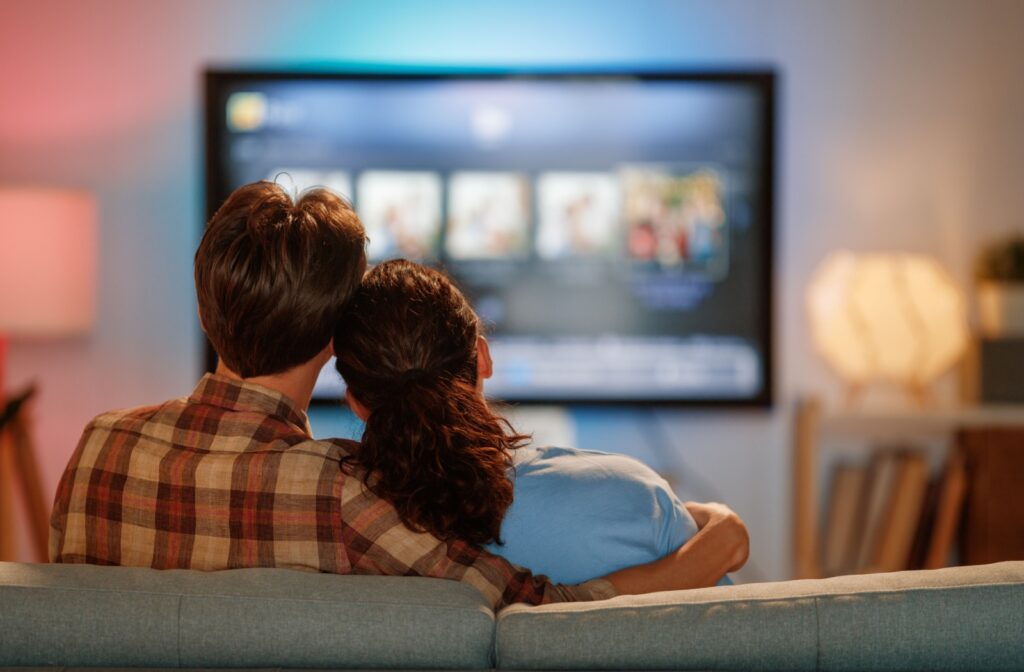 A couple sits on the couch leaning their heads together as they watch tv in the distance